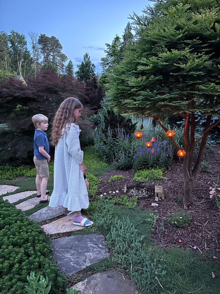 Two children looking at fairy garden with glowing lights at dusk.